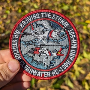 May include: A red, white, and blue embroidered patch with the text "Air Station Clearwater HC-130H and MH-60T Braving the Storm". The patch features a helicopter and a plane flying through clouds.