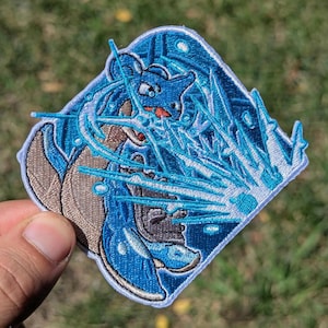 Puede incluir: Un parche bordado de forma cuadrada que presenta una criatura azul con un caparaz&oacute;n marr&oacute;n, representada en una pose din&aacute;mica mientras emite rayos de energ&iacute;a y fragmentos de color azul brillante. El dise&ntilde;o utiliza hilos de varios tonos de azul, marr&oacute;n y blanco.