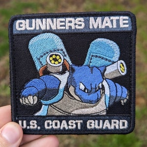May include: A black and blue embroidered patch with the text "Gunners Mate" and "U.S. Coast Guard". The patch features a cartoon blue and gray creature with a large cannon on its back.