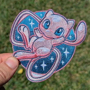 May include: An embroidered patch featuring a light pink creature with large blue eyes and a long tail, centered against a circular dark blue background filled with white four-pointed star shapes and a thin pink border outlining the entire design.