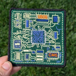 May include: A green embroidered patch with a circuit board design. The patch has the text "USCG" and "CMS" in orange.