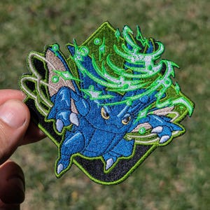 May include: An embroidered patch featuring a blue creature with yellow eyes, surrounded by swirling green energy effects and sparkles. The design is set against a black background with a green border, showcasing detailed stitching and vibrant colors.