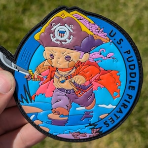 May include: A blue and black circular patch with a cartoon pirate holding a sword. The pirate is wearing a red and pink shirt, purple pants, and a yellow and brown hat with a US Coast Guard emblem. The text "U.S. PUDDLE PIRATES" is written around the edge of the patch.