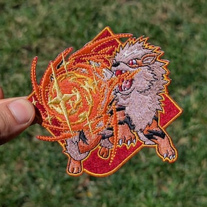 May include: An embroidered patch featuring a stylized creature with tan fur, black stripes, and red eyes, breathing a large, swirling orange and yellow fireball with star-shaped accents, set against a red, diamond-shaped background.