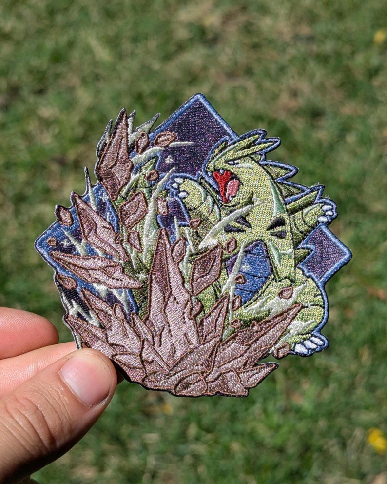 Op de afbeelding: Een geborduurde patch met een groen, tweebenig wezen met een rode mond en scherpe klauwen, geplaatst achter een cluster van bruine, grillige rotsachtige formaties tegen een donkerblauwe, ruitvormige achtergrond met een gestikte rand.