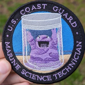 May include: A black and purple embroidered patch with the text "U.S. Coast Guard Marine Science Technician". The patch features a cartoon purple creature in a glass jar.