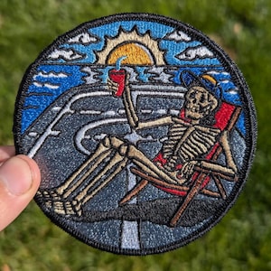 May include: Embroidered patch featuring a skeleton wearing a hat and sunglasses, relaxing in a beach chair with a drink. The background shows a beach scene with a sunset and ocean waves.