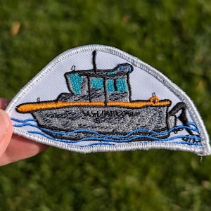 May include: A white embroidered patch with a gray tugboat with an orange stripe and blue waves. The tugboat has a blue and teal cabin.