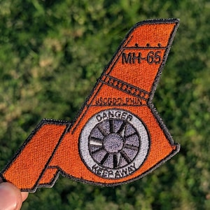 May include: An orange embroidered patch with the text "MH-65" and "USCGDOLPHIN". The patch features a circular design with the word "DANGER" and "KEEP AWAY". The patch is shaped like a tail fin.