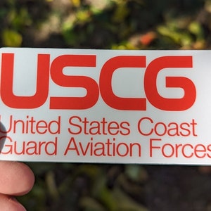 May include: A white rectangular sign with the red text "USCG" and the words "United States Coast Guard Aviation Forces" in red.