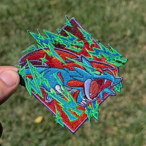 May include: An embroidered patch in a diamond shape featuring a blue creature with sharp teeth and red wings. The design is accented with bright green lightning bolt patterns and star shapes against a deep red background with a purple border.