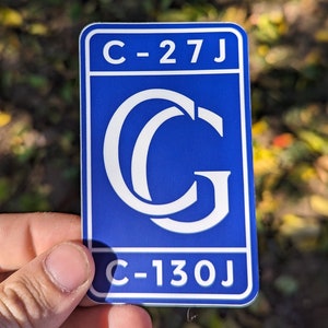 May include: Blue and white sticker with the letter "G" in a stylized font and the text "C-27J" and "C-130J".