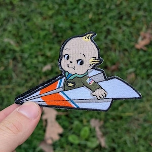 May include: Embroidered patch featuring a cartoon baby in a paper airplane. The baby is wearing a green jacket with a small patch. The paper airplane has orange and blue stripes. The patch is on a green background.
