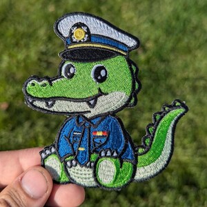 May include: A green alligator wearing a blue police officer uniform with a white and blue hat. The alligator has a friendly expression and is sitting with its tail curled behind it.