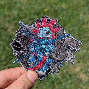 May include: An embroidered patch featuring a stylized creature with a light blue head, red crest, and dark blue body. The design includes dark gray wing-like shapes and bright blue energy lines crossing over the center of the figure.