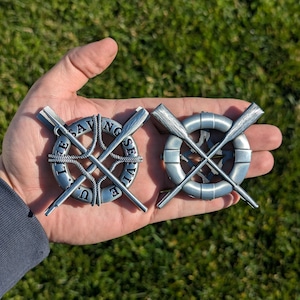 May include: Two silver-toned metal pins depicting the US Life Saving Service emblem. The emblem features a life preserver with crossed oars and the words "US Life Saving Service".