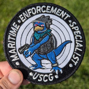 May include: Embroidered patch featuring a blue cartoon dinosaur holding a gun, with the text "MARITIME ENFORCEMENT SPECIALIST" and "USCG" in white lettering on a black background. The dinosaur wears a vest and scarf.