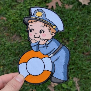 May include: Embroidered patch featuring a cartoon sailor in a blue uniform and hat, leaning on an orange and white life preserver. The patch has a detailed design with a smiling face and is held by a hand against a green background.
