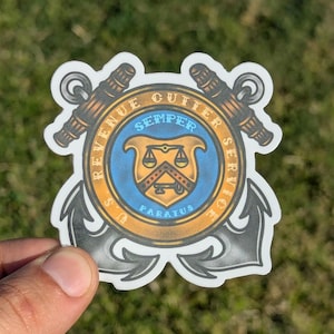 May include: A sticker featuring the US Revenue Cutter Service emblem. The design includes a central shield with scales, surrounded by text and anchors. The color scheme is primarily blue, gold, and black.