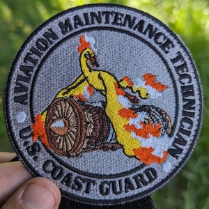 May include: A gray embroidered patch with the text "U.S. Coast Guard Aviation Maintenance Technician" in a black circle. The center of the patch features a yellow and orange stylized flame design with a brown and gray engine.