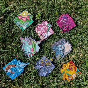 Op de afbeelding: Een verzameling van acht vierkante geborduurde patches met diverse kleurrijke wezens met dynamische actie-effecten, waaronder een groen plantwezen, een roze psychisch figuur, een paarse geest, een blauwe waterschildpad en een gele vuurdraak.