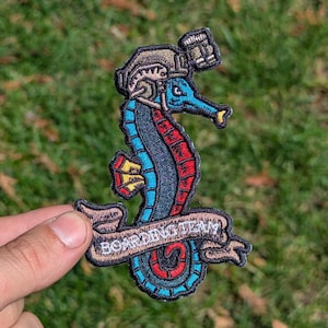 May include: Embroidered patch featuring a seahorse with a beer stein helmet and the words "BOARDING TEAM" on a banner. The seahorse is blue, red, and yellow, with a brown helmet. The patch is held against a green grassy background.