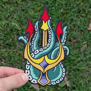 May include: Embroidered patch featuring a trident design intertwined with octopus tentacles. The patch has a color palette of teal, yellow, red, and purple, with black outlining. The patch is held against a green grassy background.