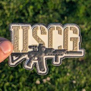 May include: A patch featuring the letters "USCG" in tan and off-white, with a black and gray rifle design beneath the letters. The patch is held against a green grassy background.