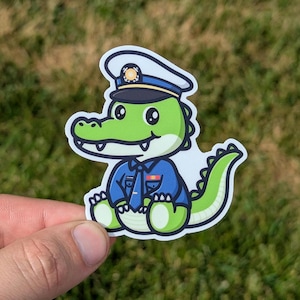 May include: A green alligator wearing a blue police officer uniform with a white hat. The alligator is sitting with its legs crossed and has a friendly expression.