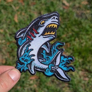 May include: Embroidered patch featuring a stylized shark with an open mouth, showcasing yellow teeth. The shark is primarily white with black and red accents, surrounded by blue waves. The patch has a black border and is likely for clothing or accessories.