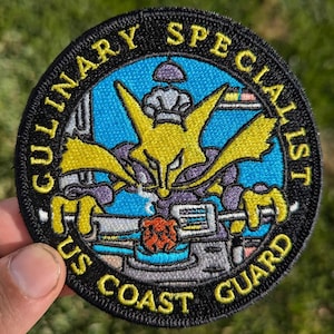 May include: A black and yellow embroidered patch with the text "Culinary Specialist US Coast Guard". The patch features a cartoon chef with a yellow body and purple clothing holding a spatula and a red meatball.