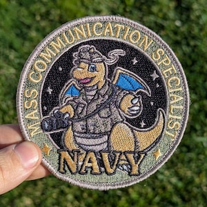 May include: A round, embroidered patch with a cartoon dragon wearing a camouflage uniform and holding a camera. The patch is beige with gold lettering that reads "Mass Communication Specialist NAVY".