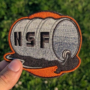 May include: Embroidered patch with the letters "NSF" in black, set against a gray barrel design with an orange and black border. The patch is likely for a uniform or bag.