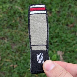 May include: A rectangular patch resembling a bullet. It has a red and white striped top, a tan body, and a black base with white text. The patch is held in a hand against a green background.