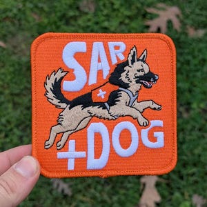 May include: An orange square patch with the text "SAR DOG" in white, depicting a running German Shepherd dog with a red cross on its vest. The patch is set against a green, grassy backdrop.