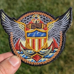 May include: A circular embroidered patch with a red, white, and blue shield with an eagle's wings on either side. The shield has a gold outline and the eagle's wings are gray. The patch has a brown rope border.