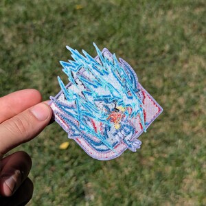 以下が含まれることがあります： ダイヤモンド型の刺繍ワッペン。青と白の龍のようなスタイリッシュな生き物が描かれ、その周囲をギザギザの稲妻のような青い刺繍が囲んでいます。明るい背景に赤と黄色の糸の小さなアクセントが施されたデザインです。