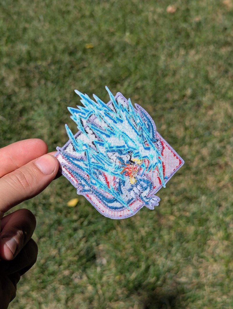 以下が含まれることがあります： ダイヤモンド型の刺繍ワッペン。青と白の龍のようなスタイリッシュな生き物が描かれ、その周囲をギザギザの稲妻のような青い刺繍が囲んでいます。明るい背景に赤と黄色の糸の小さなアクセントが施されたデザインです。