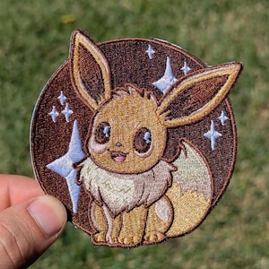 May include: A circular embroidered patch featuring a brown Eevee character with large dark eyes and a cream-colored chest ruff. The background is a dark brown circle decorated with light blue four-pointed star shapes and small white sparkles.