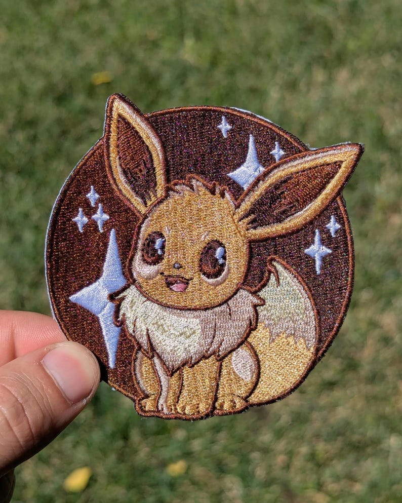 May include: A circular embroidered patch featuring a brown Eevee character with large dark eyes and a cream-colored chest ruff. The background is a dark brown circle decorated with light blue four-pointed star shapes and small white sparkles.