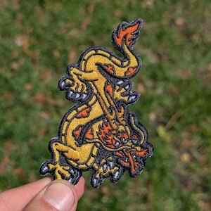May include: Embroidered patch featuring a stylized yellow dragon with orange and black accents. The dragon has a detailed design with a black outline. The patch is held against a blurred green background.
