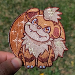 May include: A circular embroidered patch featuring a stylized brown and cream-colored creature with large eyes, pointed ears, and a fluffy mane. The background of the patch is a deep orange circle decorated with several four-pointed star shapes.