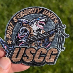 May include: Embroidered patch featuring a shark-soldier with a machine gun. The patch reads "PORT SECURITY UNIT" and "USCG" in a circular design. The shark is in shades of blue, brown, and pink, with a menacing expression. The patch is held by a hand.