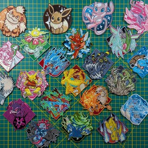 May include: A collection of twenty-two embroidered patches featuring various colorful cartoon creatures in dynamic poses. Each patch displays a unique character, including fire, water, and psychic types, rendered with detailed stitching on a green grid.