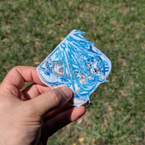 Puede incluir: Un parche bordado que presenta un personaje de anime estilizado con cabello largo y azul fluido y detalles intrincados en hilo blanco y azul. El parche tiene forma de diamante con un borde blanco y costuras detalladas en todo el dise&ntilde;o.