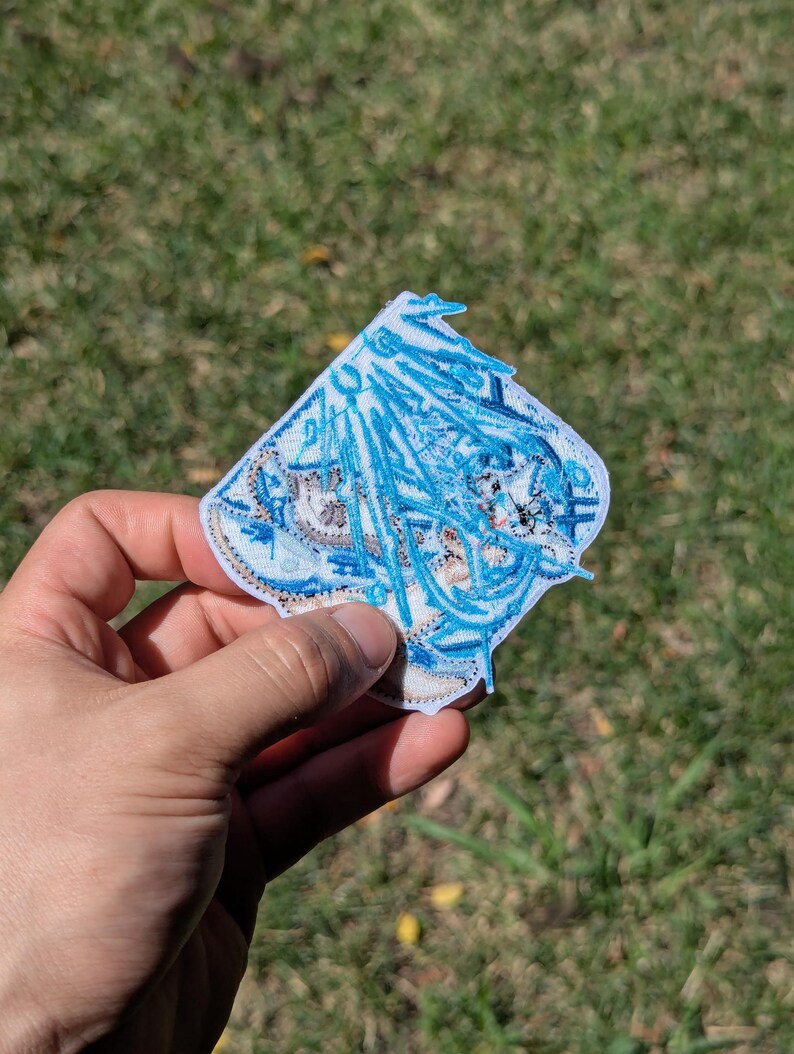 Puede incluir: Un parche bordado que presenta un personaje de anime estilizado con cabello largo y azul fluido y detalles intrincados en hilo blanco y azul. El parche tiene forma de diamante con un borde blanco y costuras detalladas en todo el dise&ntilde;o.