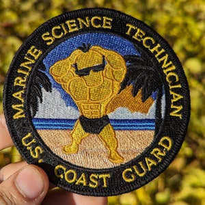 May include: A black and yellow embroidered patch with the text "U.S. Coast Guard Marine Science Technician". The patch features a cartoon of a muscular person wearing sunglasses and a black swimsuit standing on a beach with palm trees.