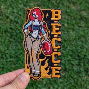 May include: Embroidered patch featuring a stylized woman with red hair, tattoos, and work attire, holding a red helmet. The patch has the word "BECCE" in orange block letters on a black background with flames.