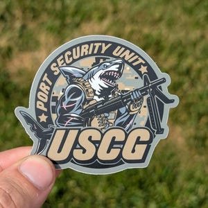 Peut inclure: Un autocollant rond avec un fond de camouflage représentant un requin de dessin animé tenant une mitrailleuse. Le texte "PORT SECURITY UNIT" est écrit autour du haut de l'autocollant et "USCG" est écrit en grandes lettres en bas.