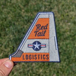 May include: A triangular embroidered patch shaped like an aircraft tail fin. The design features orange and light gray sections with the text Red Tail and LOGISTICS in dark blue. A central circular emblem displays a white star with blue wings.